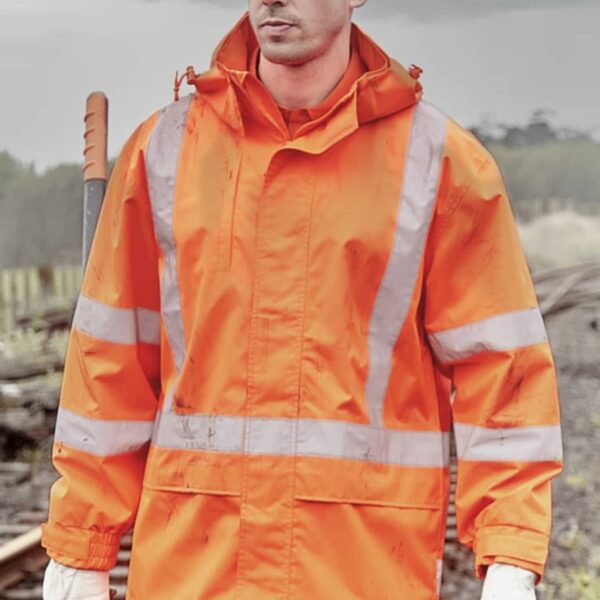 A person wearing a white hard hat, an orange high-visibility jacket with reflective stripes, and white gloves, standing near train tracks. They are holding a tool and looking to the side, with a cloudy sky in the background.