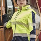 A person wearing a bright yellow safety jacket and a white hard hat stands leaning against a construction vehicle, looking into the distance. The setting appears to be a construction site.