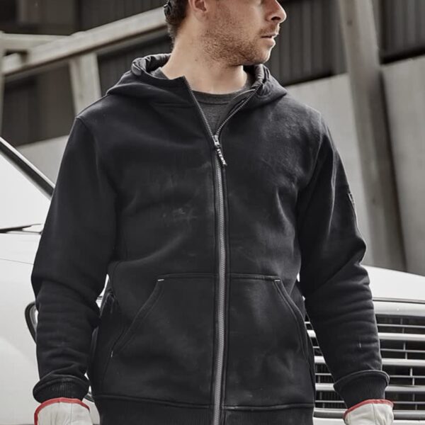 A construction worker wearing a white hard hat, black hoodie, and white gloves stands in front of a vehicle. The background features industrial equipment.