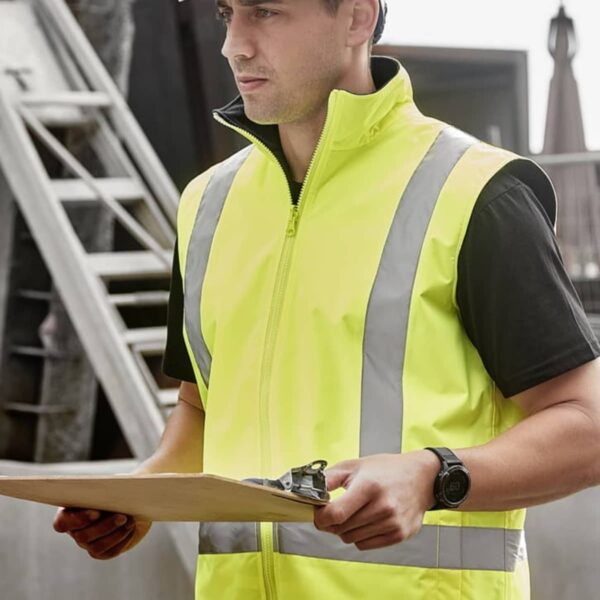 A construction worker wearing a white hard hat and a bright yellow safety vest holds a clipboard. He stands in an outdoor setting with construction materials and a ladder in the background.
