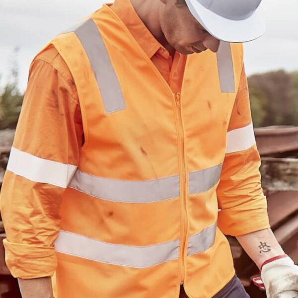 A person wearing a white hard hat and an orange reflective safety vest holds a tool. They are looking down, with trees and metal beams in the background.