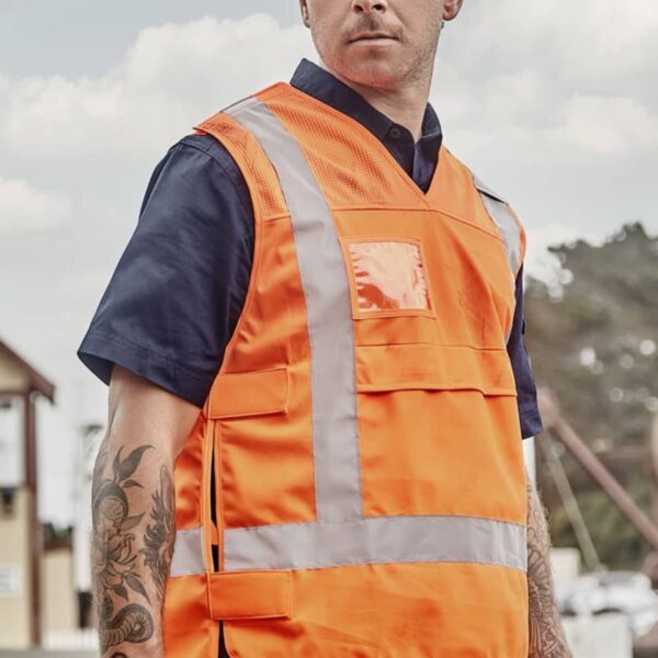 A man wearing a bright orange safety vest with reflective stripes stands outdoors. He has a navy blue shirt underneath and has tattoos on his left forearm. The background shows a cloudy sky and partial buildings.