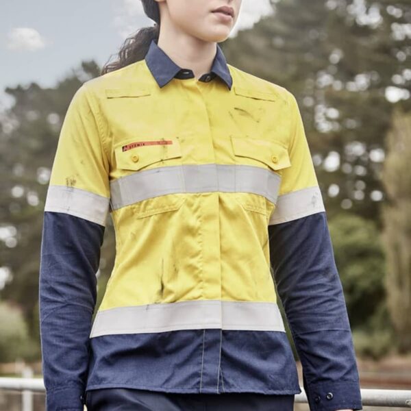 A person wearing a yellow and navy blue high-visibility work shirt and a white hard hat stands outdoors. They are holding gloves and looking to the side, with trees and a cloudy sky in the background.