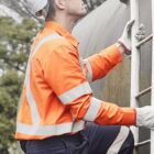 A construction worker wearing an orange safety jacket, white hard hat, and gloves climbs a metal ladder outdoors. He looks upward, next to a large industrial pipe, with trees in the background.