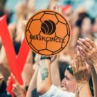 A large crowd of people raising their hands and cheering at an event. A large orange soccer ball-shaped logo with the text MASKCIRCLE is overlaid prominently in the center of the image.