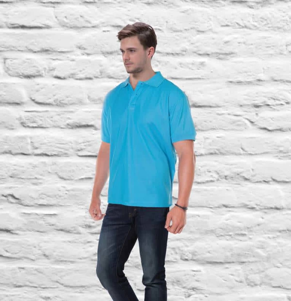 Wearing the AERO POLO in vibrant blue and dark jeans, a man stands against a white brick backdrop. He gazes to the side, with one hand in his pocket as he steps slightly forward.