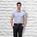 A man wearing a Euro POLO in light gray and black pants stands with hands in pockets against a white brick wall. He has dark hair and a slight smile.