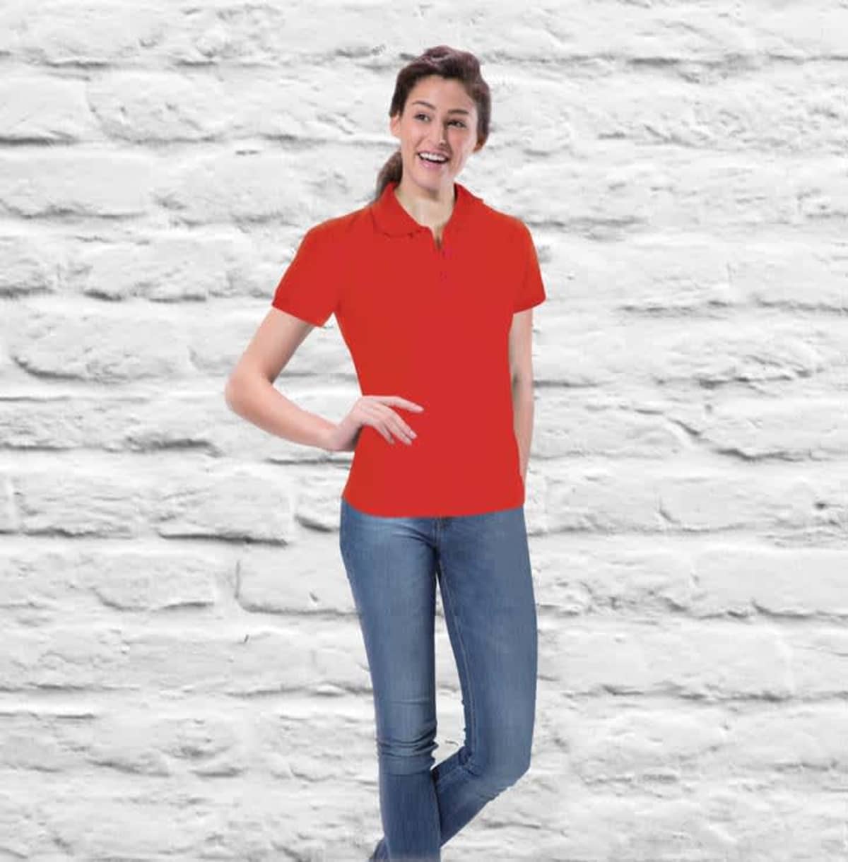 Wearing a Velocity Ladies Polo with blue jeans, a person leans against a white brick wall with casual confidence. A smile highlights their effortless style and charm as one hand rests on their hip.