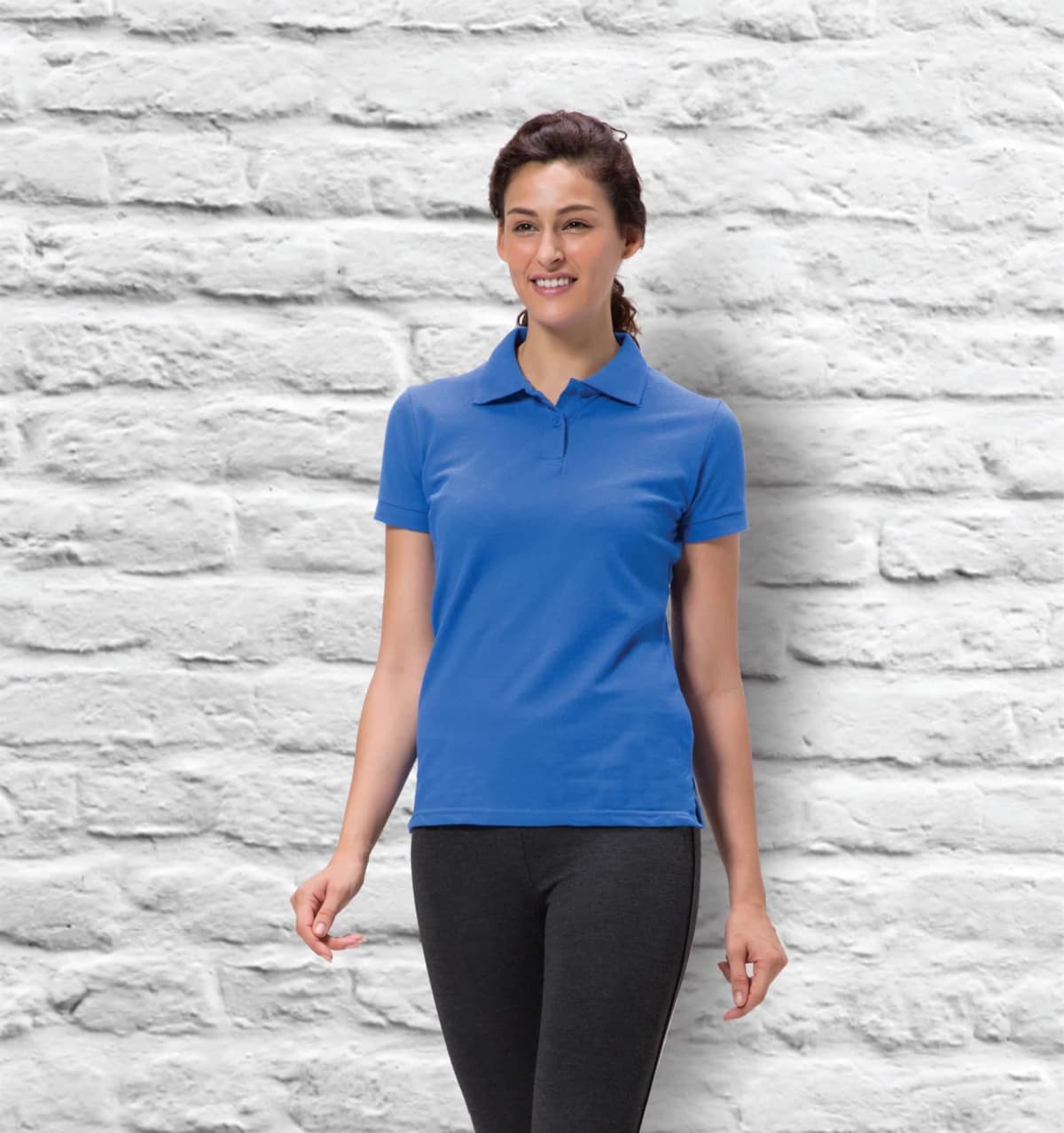 A woman in a blue Heritage Ladies Polo and dark pants smiles confidently in front of a white brick wall, subtly embodying timeless heritage.