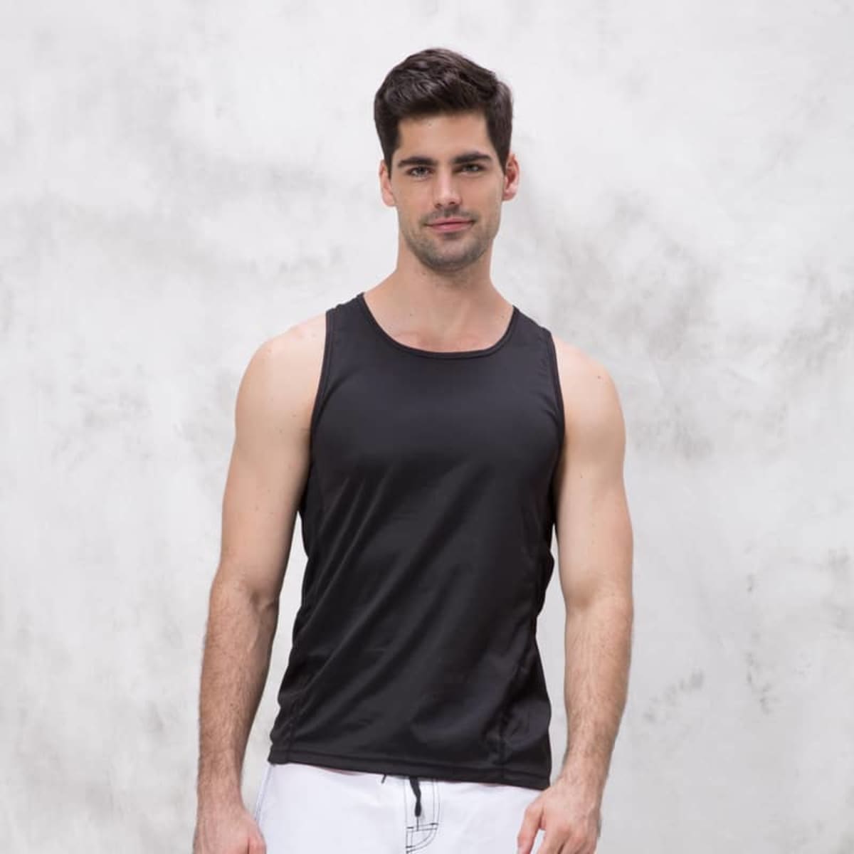 A man in a Triumph Singlet and white shorts stands against a textured light gray background, looking directly at the camera with a slight smile.