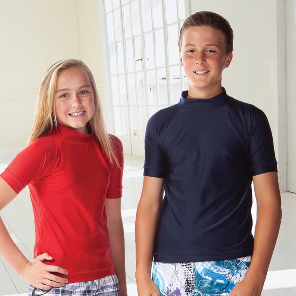 Near a bright window, two children stand indoors. The girl on the left wears a red shirt and checkered shorts, smiling at the boy on the right who wears a Shield Rashie and promotional-patterned shorts. Their joyful expressions light up the room as they stand side by side.
