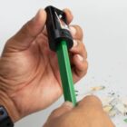 A hand sharpening a green pencil with a black sharpener; pencil shavings are visible.
