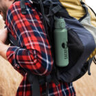 Person in plaid shirt hiking with a backpack and a green Sherpa water bottle attached.