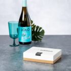 A bottle of Lolo wine, a teal glass, a leaf, and a white box on a grey surface.