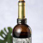Brown glass wine bottle with a gold neck and a patterned label, in front of a leafy background.
