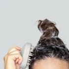 Person using a scalp brush to wash hair with shampoo and foam against a gray background.