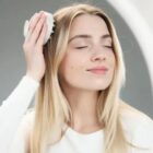 Woman with closed eyes brushing her long blonde hair with a white brush, looking relaxed.
