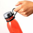 A hand holding a red water bottle by its black loop near the silver cap.