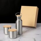 Metal bottle and two cups with wooden lids, placed in front of a plain cardboard box.