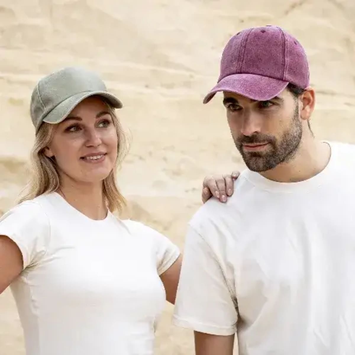 A woman and man in white shirts and baseball caps stand together against a sandy background.