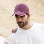Man in a white shirt and purple cap standing in front of a beige wall.