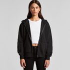 Woman in black zip-up hoodie and white crop top, standing against a plain gray background.