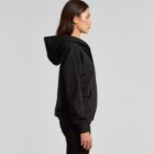 Woman in a black hoodie and pants standing in profile against a plain light gray background.