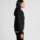 Woman in a black hoodie and pants standing sideways against a plain light background.