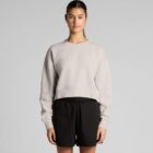 Woman in a beige sweater and black shorts standing against a plain gray background.