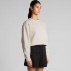Woman in beige sweatshirt and black shorts standing against a plain gray background.