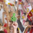 A crowd of people enthusiastically cheering at an event, with raised hands and vibrant expressions. In the foreground, one person holds up a green hand-shaped foam sign with a thumb up.