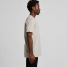Man in a beige t-shirt and black pants standing in profile against a plain background.