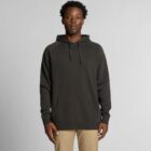 Man wearing a black hoodie and khaki pants standing against a plain gray background.