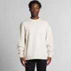A man wearing a cream-colored sweatshirt stands against a plain light gray background.