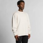 Man wearing a cream-colored sweatshirt and black pants, standing against a plain background.