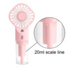 A pink handheld fan with a transparent detachable water tank. The tank has a marked 20ml scale line. The fan features a circular grid design and a power button below the head. A logo is visible on the front.