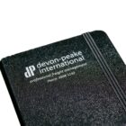 A black notebook with devon.peake international and professional freight management printed on the cover in white text. It includes a phone number and has an elastic band for closure.