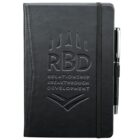 Black notebook with RBD and Relationship Breakthrough Development embossed on the cover. Features an elastic band closure and a pen attached to the side in a loop.