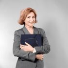 Smiling woman with short, light brown hair, wearing a gray suit, holds blue folders against a light gray background.