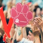 A crowd of people cheering with their hands raised, holding red cheering sticks and a large foam paw with the word COMPANY on it. The scene conveys excitement and support at an outdoor event.