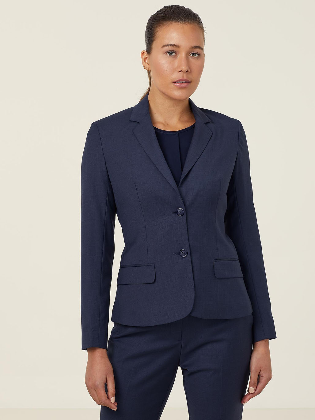 A person wearing a fitted navy blue suit with a blazer and matching pants stands against a plain light background. The blazer has two buttons and flap pockets. The person has long hair tied back and appears to be looking towards the camera.