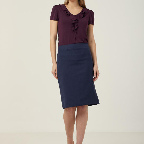 A person stands smiling against a plain background, wearing a burgundy ruffled blouse, a dark blue knee-length skirt, and brown heels. They have long, straight hair and are looking slightly to the side.