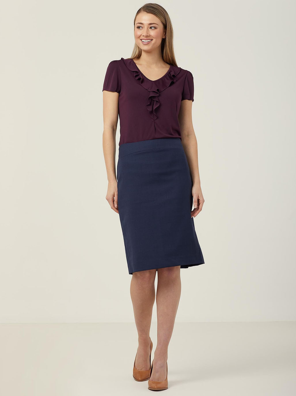 A person stands smiling against a plain background, wearing a burgundy ruffled blouse, a dark blue knee-length skirt, and brown heels. They have long, straight hair and are looking slightly to the side.