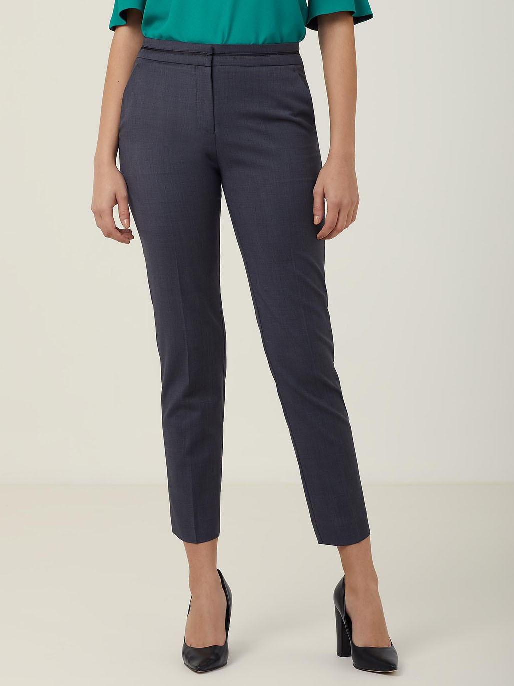 A person wearing teal green blouse and dark gray tailored pants, standing against a plain light background. Their feet are in black high heels. The image is focused on the lower body.