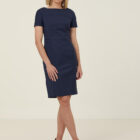A woman in a navy knee-length dress and tan heels, standing against a plain light background. She has shoulder-length blonde hair and is posing with one leg slightly crossed in front of the other, with a neutral expression.