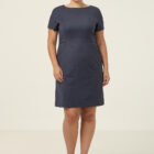 A person stands against a plain background, wearing a dark blue, short-sleeved dress and black heels. They are smiling with their hands by their sides.