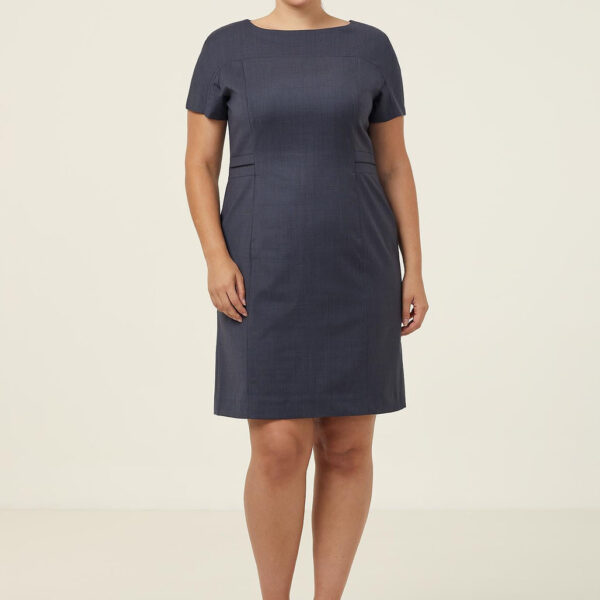 A person stands against a plain background, wearing a dark blue, short-sleeved dress and black heels. They are smiling with their hands by their sides.