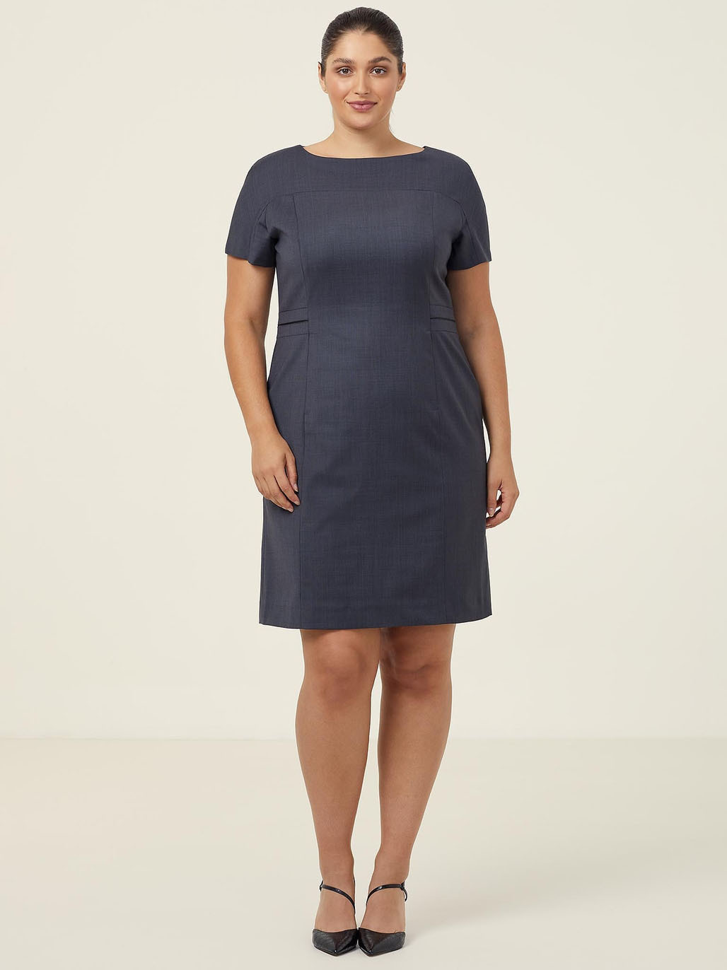 A person stands against a plain background, wearing a dark blue, short-sleeved dress and black heels. They are smiling with their hands by their sides.