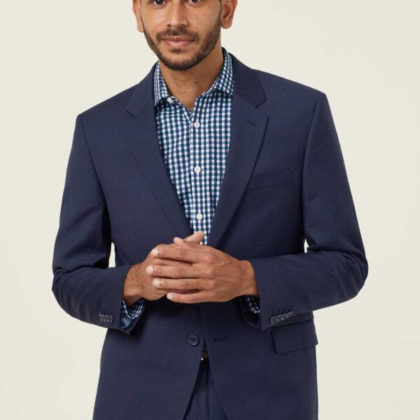 A man with short dark hair and a beard is wearing a navy suit jacket over a blue and white checkered shirt. He stands against a plain light background, looking slightly to the side with his hands gently clasped together.