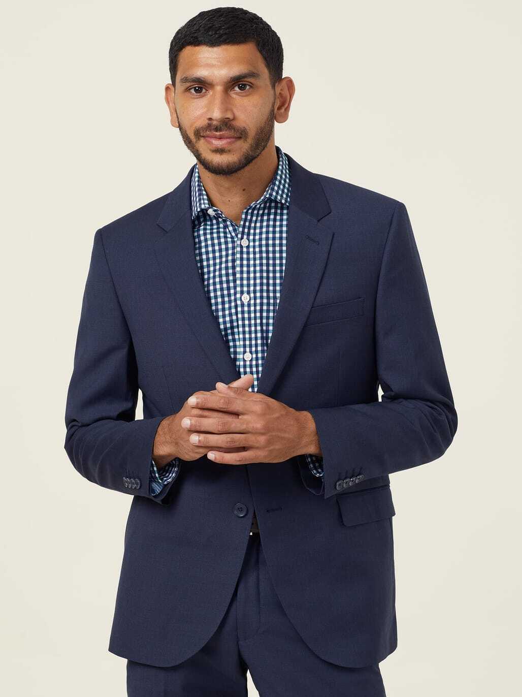 A man with short dark hair and a beard is wearing a navy suit jacket over a blue and white checkered shirt. He stands against a plain light background, looking slightly to the side with his hands gently clasped together.